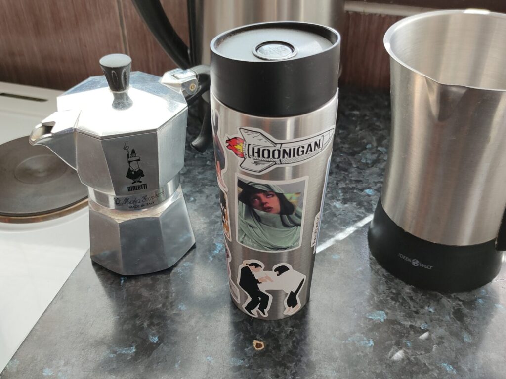 An image of a stainless steel coffee tumbler with a moka pot to the left and a milk frother to the right.